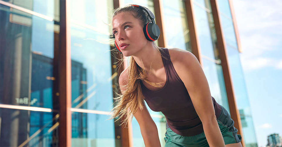 Vrouw met Philips A6219-koptelefoon neemt een pauze tijdens een training in de buitenlucht, staand voor een modern glazen gebouw