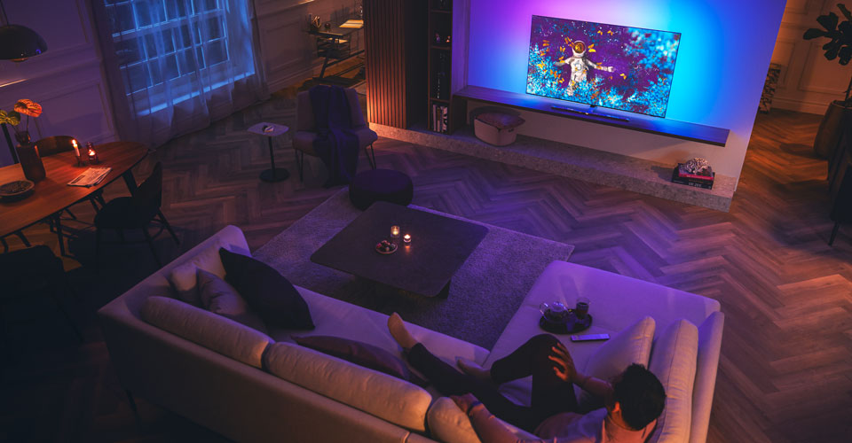 Man ontspant op een bank in een schemerige woonkamer en kijkt naar een Philips Ambilight-tv